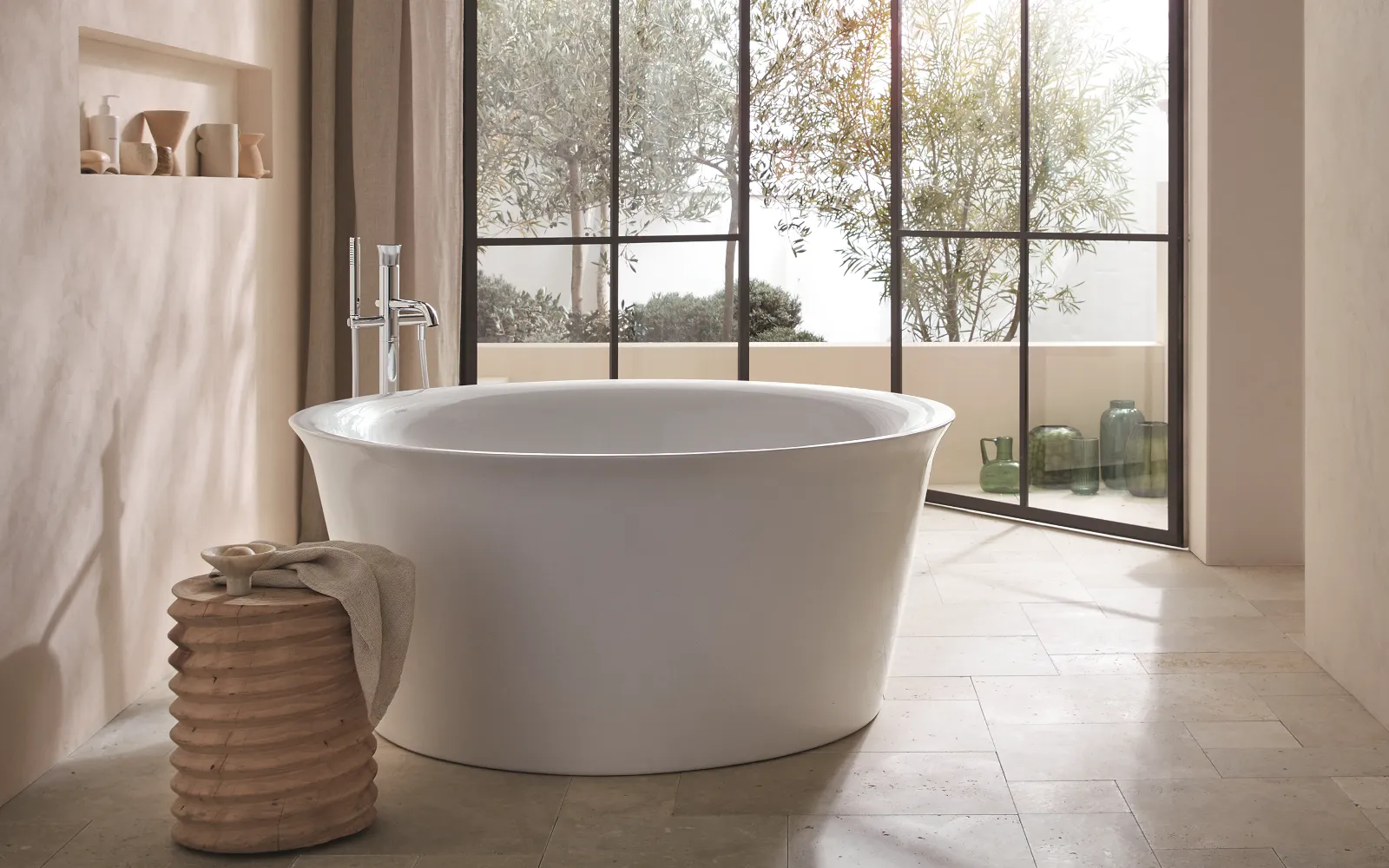 Ein luxuriöses Badezimmer mit einer großen, freistehenden Badewanne vor bodentiefen Fenstern, die auf eine Terrasse mit Pflanzen und grünen Glasvasen blicken. 