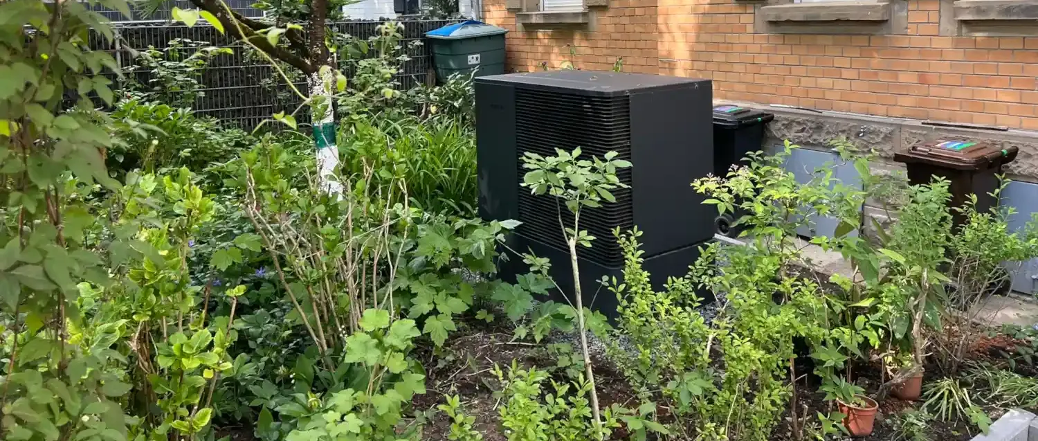 Eine moderne Luft-Wasser-Wärmepumpe ist in einem dicht begrünten Vorgarten vor einem Mehrfamilienhaus mit Klinkerfassade installiert. Das anthrazitfarbene Außengerät fügt sich harmonisch zwischen Sträuchern, Bäumen und Blumenbeeten ein und sorgt für eine nachhaltige Wärmeversorgung. Die unauffällige Platzierung verbindet hohe Energieeffizienz mit einer ansprechenden Gartengestaltung.