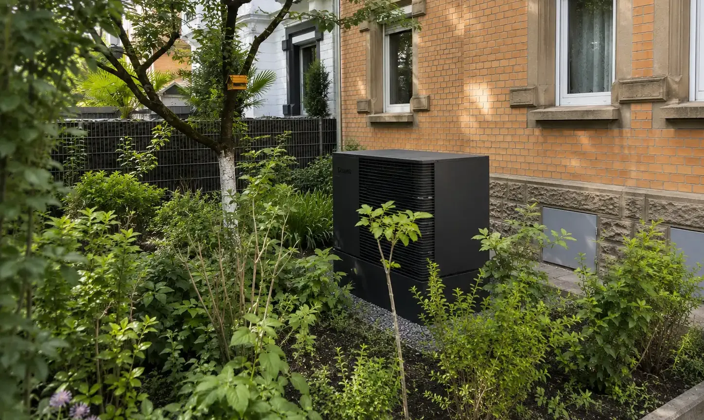 Eine kompakte Wärmepumpen-Außeneinheit befindet sich in einem begrünten Vorgarten vor einem Mehrfamilienhaus und ist von Hecken, Bäumen sowie Zierpflanzen umgeben. Das schlichte anthrazitfarbene Design passt sich unauffällig an die Umgebung an und ermöglicht eine effiziente Nutzung erneuerbarer Energie. Die fachgerechte Installation verbindet Nachhaltigkeit mit einer optisch gelungenen Integration.