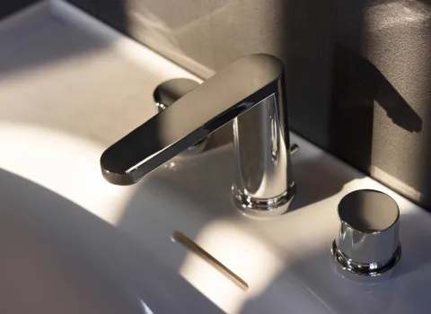 Ein moderner, verchromter Wasserhahn mit flachem Auslauf ist auf einem weißen Waschbecken montiert.