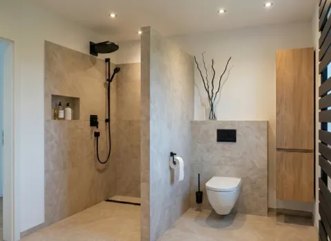 Badezimmer mit begehbarer Dusche in Natursteinoptik, schwarzer Regendusche und stilvollem Hänge-WC neben Holzschrank.