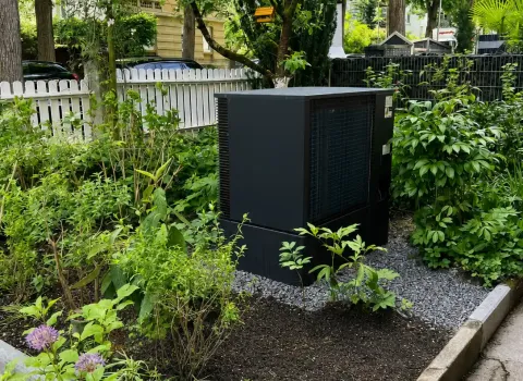 Eine kompakte Wärmepumpen-Außeneinheit befindet sich in einem liebevoll angelegten Vorgarten mit Bäumen, Sträuchern und blühenden Pflanzen. Das schlichte anthrazitfarbene Design passt sich dezent an die grüne Umgebung an und sorgt für eine nachhaltige Wärmeversorgung des Gebäudes. Die professionelle Installation verbindet hohe Energieeffizienz mit einer optisch gelungenen Einbindung in die Außenanlage.