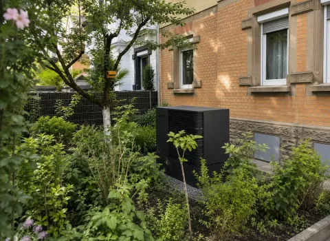 Eine kompakte Wärmepumpen-Außeneinheit befindet sich in einem begrünten Vorgarten vor einem Mehrfamilienhaus und ist von Hecken, Bäumen sowie Zierpflanzen umgeben. Das schlichte anthrazitfarbene Design passt sich unauffällig an die Umgebung an und ermöglicht eine effiziente Nutzung erneuerbarer Energie. Die fachgerechte Installation verbindet Nachhaltigkeit mit einer optisch gelungenen Integration.