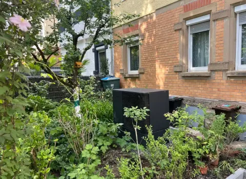 Eine kompakte Wärmepumpen-Außeneinheit befindet sich in einem begrünten Vorgarten vor einem Mehrfamilienhaus und ist von Hecken, Bäumen sowie Zierpflanzen umgeben. Das schlichte anthrazitfarbene Design passt sich unauffällig an die Umgebung an und ermöglicht eine effiziente Nutzung erneuerbarer Energie. Die fachgerechte Installation verbindet Nachhaltigkeit mit einer optisch gelungenen Integration.
