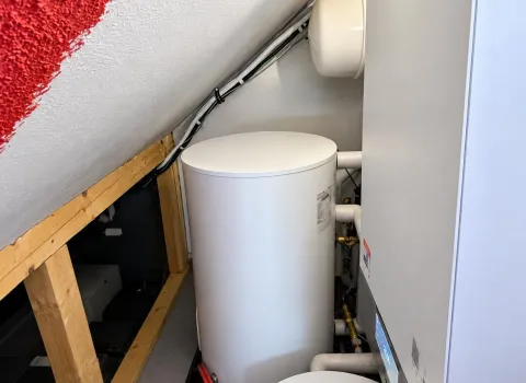 Moderne Heizungsanlage mit Wärmepumpe und Warmwasserspeicher, platzsparend installiert in einem Technikraum unter einer Dachschräge.