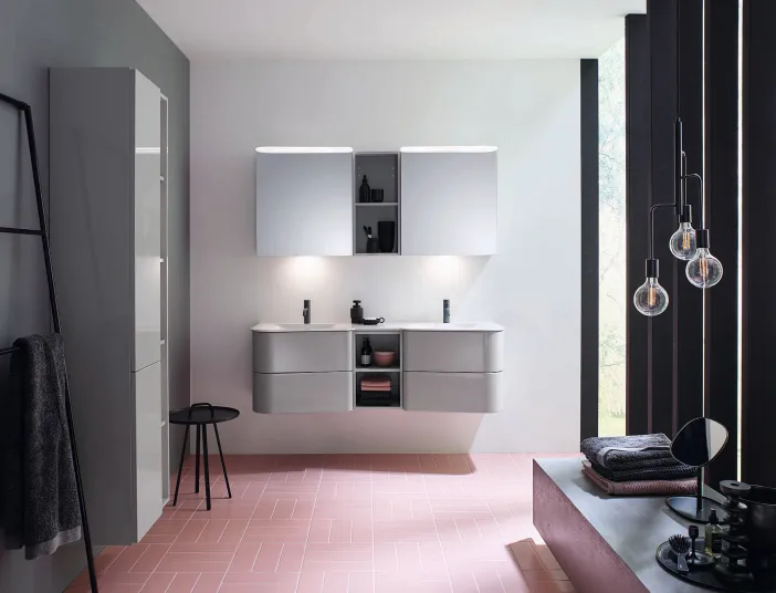 Modernes Badezimmer mit zwei Lichtspiegelschränken, eleganten grauen Schränken, rosafarbenen Fliesen und hängenden Pendeleuchten.