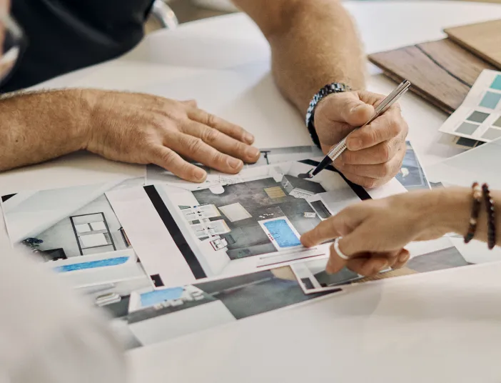 Zwei Personen besprechen ein Badezimmer-Projekt und wählen Farben aus einer Palette. Hände zeigen auf einen Grundriss mit freistehennder Wanne und Badezimmer-Möbeln.